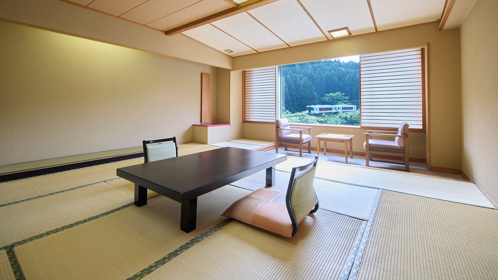 昼の光が入る鳴子温泉ホテル亀屋の東館・和室12.5畳の客室。開けられた障子窓の向こうをJR陸羽東線の列車が走行しており、手前には長方形の座卓と座椅子が置かれている